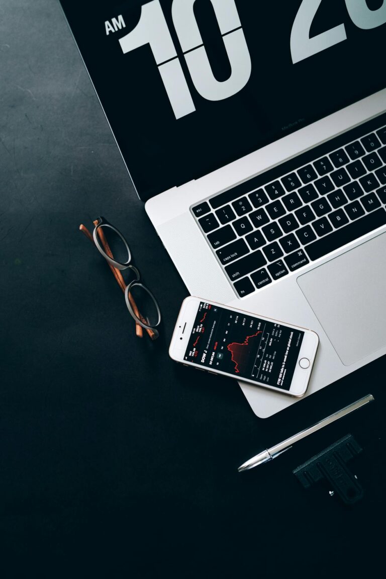 A modern workspace featuring a laptop, smartphone with stock data, and eyeglasses.