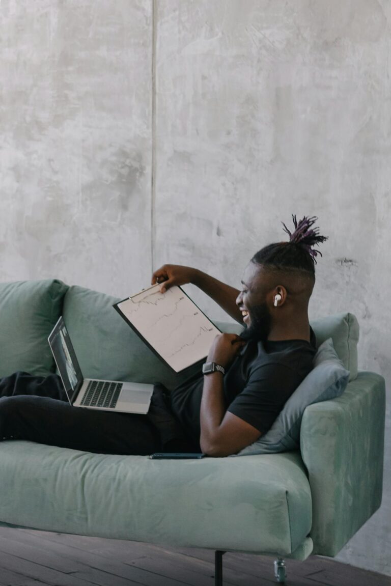 Bearded man working on a laptop while reclining on a sofa, analyzing data on a clipboard.