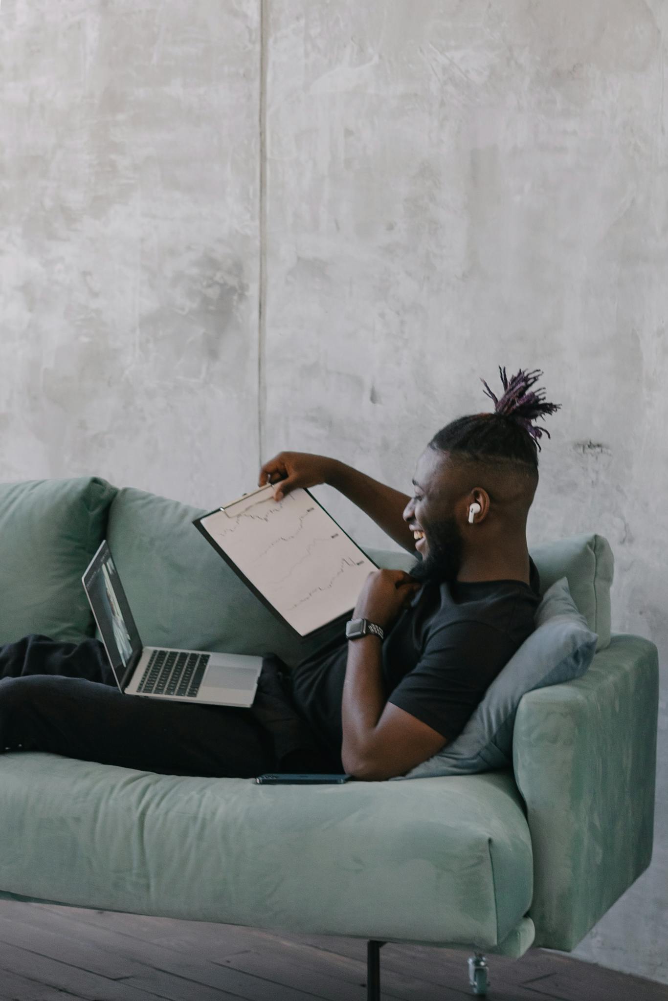 Bearded man working on a laptop while reclining on a sofa, analyzing data on a clipboard.